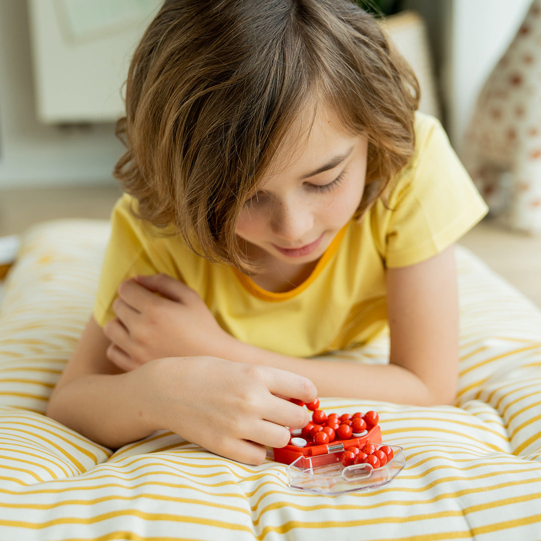 Photo of child lying on a yellow and white striped cushion playing IQ Mini Hexpert mind puzzle by Smart Games.